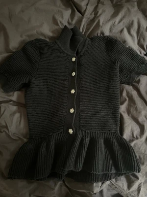 Svart stickad topp med volang - Säljer en svart stickad topp med korta ärmar, hög krage och volang nedtill. Knäpps framtill med blommiga, glittriga knappar. Materialet är ribbstickat. Knappt använd och kanske har lite damm men går lätt att tvätta bort!
