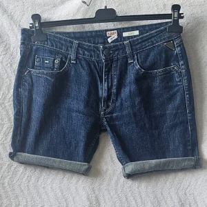 Blå denimshorts från Replay - Snygga blå denimshorts från Replay med klassisk femficksdesign och uppvikta benslut. Shortsen har normal passform, knappgylf och detaljerade sömmar. Perfekta för en avslappnad stil under varmare dagar.
