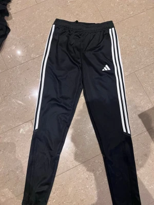 Svarta Adidas träningsbyxor - Säljer ett par svarta träningsbyxor från Adidas med klassiska vita ränder längs sidorna och Adidas-logga på låret. Byxorna har resår i midjan och är tillverkade i ett lätt och stretchigt material, perfekta för träning eller chill.