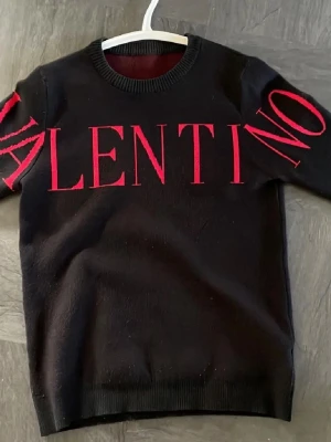 Svart stickad tröja från Valentino - Snygg svart stickad tröja från Valentino med stora röda bokstäver över bröstet och armarna. Tröjan har rund halsringning och långa ärmar. Perfekt för dig som gillar statement-plagg och exklusiv design.