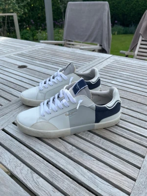 Pepe Jeans skor - Snygga sneakers från Pepe Jeans i vitt och marinblått med grå detaljer. Har bara använt skorna ett par gånger. 9/10 skick. Storlek 41, 7Eu.