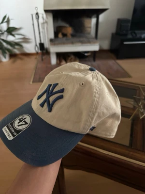 Beige och blå New York Yankees keps - Snygg beige keps från 47 Brand med mörkblå skärm och broderad New York Yankees-logga framtill. Justerbart spänne bak för perfekt passform. Klassisk baseballstil i bomull som passar till de flesta outfits.