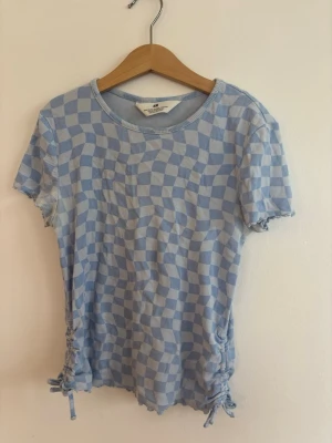 Blå och vit rutig topp med snörning - Blå och vit rutig topp från H&M med korta ärmar och snörningsdetaljer i sidorna. Toppen har en rund halsringning och är tillverkad i ett mjukt, ribbat material som ger en skön och snygg passform.