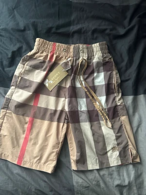 Rutig Burberry shorts med snörning - Säljer ett par klassiska Burberry shorts med det ikoniska rutiga mönstret i beige, brunt, svart och rött. Shortsen har elastisk midja med snörning och är tillverkade i ett lätt och bekvämt material, perfekta för sommaren. Inget på bilden påverkar shortsen, dem är lite sönder men det gör inget man ser inget igenom elle något alls 