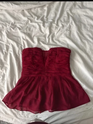 Vinröd axelbandslös topp med volang - Snygg vinröd axelbandslös topp med volang nedtill och draperad överdel. Toppen har bandeau-modell. Aldrig använd