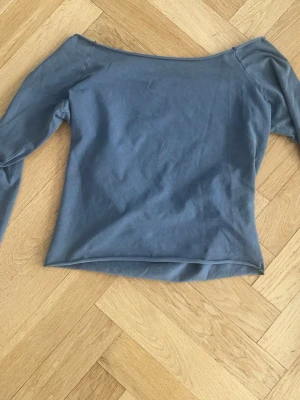 Blå off shoulder topp med lång ärm - Snygg blå off shoulder topp med långa ärmar och figurnära passform. Materialet är mjukt och stretchigt, perfekt för att skapa en cool och avslappnad look. Enkel design utan mönster eller logga, passar dig som gillar stilrena plagg.