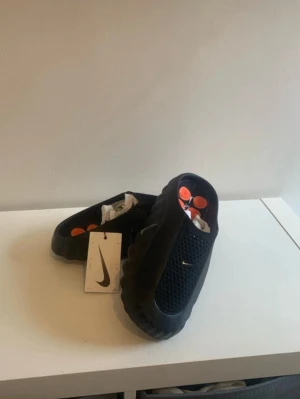 Svarta Nike Mules tofflor - Säljer ett par svarta Nike Mules tofflor med meshdetaljer och orange innersula. Tofflorna har en chunky sula och en modern, sportig look. Perfekta för dig som gillar streetwear och vill ha något bekvämt att glida runt i hemma eller ute. Kopior, kommer utan box