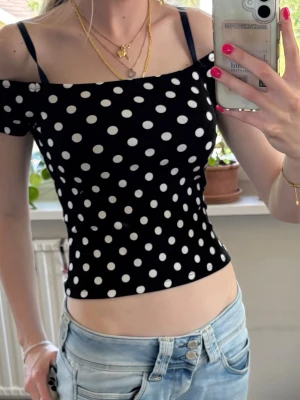 Svart prickig topp - Snygg svart croppad topp med vita prickar och offshoulder-design. Toppen har smala axelband och kort ärm, vilket ger en trendig och ungdomlig look. Perfekt att matcha med jeans eller kjol för en cool stil. Super skön men tyvärr aldrig använd💕💕💕