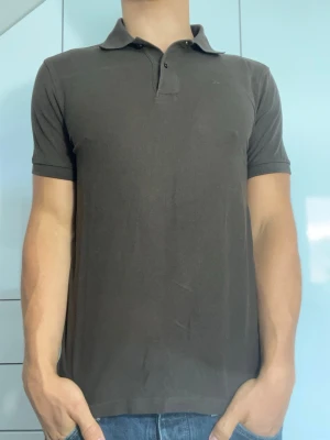Mörkbrun piké från J.Lindberg - Mörkbrun piké från J.Lindberg i 100% bomull. Strl S. Lite missfärgad vid axeln (se bild 4). Modellen är 178 cm och väger 77 kg. Hör gärna av dig vid frågor!
