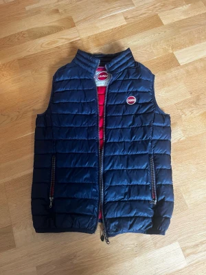 Mörkblå dunväst från Colmar - Snygg mörkblå dunväst från Colmar med quiltad design och hög krage. Västen har två dragkedjefickor framtill och röd insida som ger en cool kontrast. Colmar-logga på bröstet och dragkedja hela vägen. Perfekt för lager-på-lager och streetstyle.