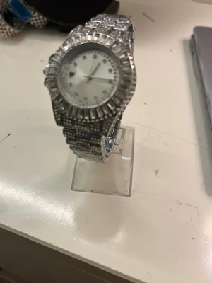 Glittrig silverfärgad klocka - Säljer en riktigt glittrig klocka i silverfärg med massor av stenar runt urtavlan och på armbandet. Klockan har en rund form och metallarmband som ger en lyxig känsla. Perfekt för dig som vill sticka ut och gillar bling!