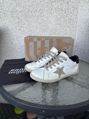 Golden Goose vita Superstar sneakers - Golden Goose Superstar sneakers i vitt läder med grå stjärna på sidan och svarta detaljer vid hälen. Skorna har beige snören och en klassisk låg siluett. Snygg och avslappnad design som passar till många olika stilar. Hör gärna av er om byten eller funderingar! Vet ej om äkthet därav priset!!