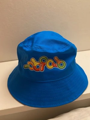 Blå bucket hat från Haribo - Cool blå bucket hat från Haribo med färgglatt godismotiv framtill och Haribo-logga på sidan. Hatten är gjord i ett lätt tygmaterial och har en klassisk form med bred skärm. Perfekt för dig som gillar streetwear och vill sticka ut med en unik accessoar.