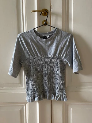 Grå ribbad topp med kort ärm - Säljer en ljusgrå topp från H&M med ribbad, smockad midja och korta ärmar. Toppen har rund halsringning och är gjord i mjukt bomullsmaterial. Perfekt för en avslappnad och trendig look.