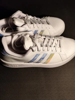 Vita Adidas sneakers med pastellränder - Snygga vita sneakers från Adidas med klassiska tre ränder i pastellfärger – blått, gult och rosa. Skorna har lågt skaft, vit sula och Adidas-logga på hälen. Tillverkade i syntetmaterial och textil för en fräsch och modern look.