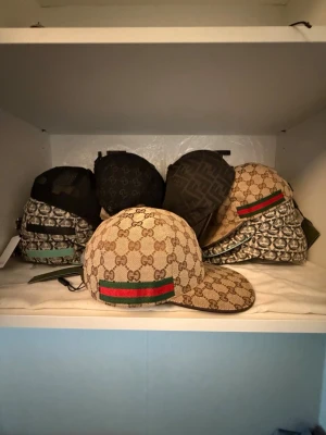 Gucci beige keps med GG-mönster - Säljer en beige keps från Gucci med det klassiska GG-monogrammet över hela kepsen och en grön-röd rand på sidan. Kepsen har böjd skärm och justerbar rem bak. Materialet är canvas med detaljer i läder. Perfekt för dig som vill ha en exklusiv och trendig accessoar.