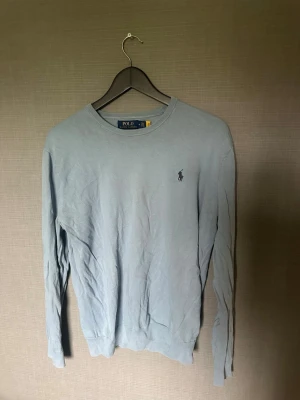Ljusblå tröja från Polo Ralph Lauren - Ljusblå långärmad tröja från Polo Ralph Lauren med rund halsringning och den klassiska broderade loggan på bröstet. Tröjan har ribbade muddar vid ärmslut och nederkant. Perfekt för en clean och stilren look.