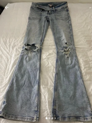 Ljusblå slitna bootcut jeans Gina Tricot - Säljer ett par ljusblå bootcut jeans från Gina Tricot i storlek XS. Jeansen har slitna detaljer och stora hål vid knäna för en trendig look. Klassisk femficksmodell i mjuk denim med snygg tvätt. Perfekta för dig som gillar en avslappnad och edgy stil.