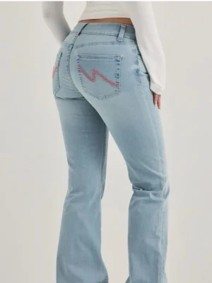 Ljusblå bootcut jeans med zigzag-detalj - Snygga ljusblå bootcut jeans med rosa zigzag-broderi på bakfickorna. Jeansen har hög midja, klassisk femficksdesign och raka ben som går ut lite nedtill. Perfekta för en trendig och avslappnad look.