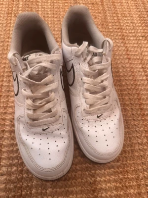 Vita Nike Air Force 1 sneakers - Klassiska vita Nike Air Force 1 sneakers med svart swoosh-logga på sidan och AIR-text bak. Skorna har perforerad tå för ventilation, vita skosnören och är tillverkade i läder. Perfekt för dig som gillar en clean och tidlös stil.