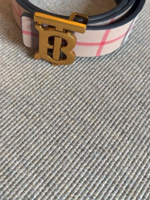 Burberry bälte med guldspänne 115cm - Snyggt Burberry bälte med klassiskt rutigt mönster i beige, rosa och grått. Bältet har ett stort guldigt B-spänne och kommer med originalpåse, ask och Burberry-kort. Perfekt accessoar för att lyfta din outfit.