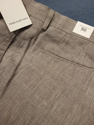 Beige linnebyxor These Glory Days 50 M - Snygga kostymbyxor från These Glory Days i beige och grått rutmönster. Klassisk rak passform med bälteshällor, knapp och dragkedja. Tunt, vävt material som ger en stilren look. Perfekta för dig som vill ha en modern twist på klassiska kostymbyxor.