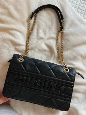 Valentino Axelväska  - Snygg svart quiltad handväska från Valentino med guldiga kedjehandtag och stort logotyptryck på framsidan. Väskan har en klassisk rektangulär form och är tillverkad i vadderat konstläder. Perfekt accessoar för att lyfta din outfit.