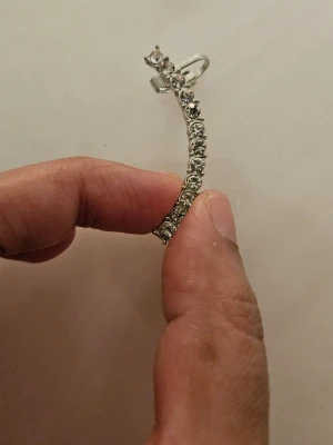 Silvriga örhängen med gnistrande stenar - Säljer ett par silvriga örhängen med rad av gnistrande, runda stenar. Örhängena har en böjd form och stift med säkerhetslås. Perfekta för dig som gillar stilrena och glittriga accessoarer. Inget par. Har aldrig användts. Har allergi. 