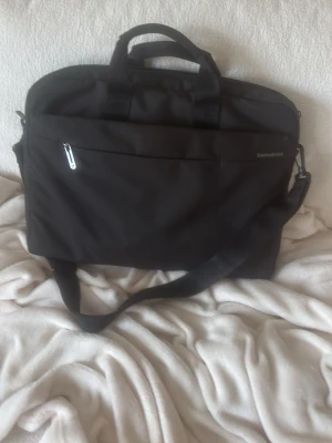 Svart datorväska från Samsonite - Snygg svart datorväska från Samsonite med justerbar axelrem och två bärhandtag. Väskan har en stor ytterficka med dragkedja och är tillverkad i slitstarkt tygmaterial. Perfekt för laptop och tillbehör när du är på språng.