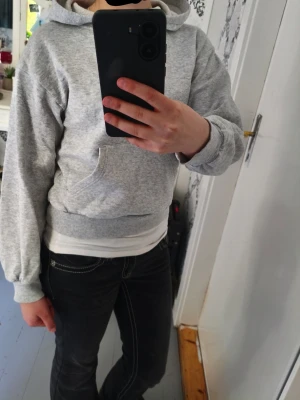 Ljusgrå hoodie med huva - Säljer en enkel och stilren ljusgrå hoodie med huva och magficka. Hoodien har ribbade muddar vid ärmslut och nederkant samt en avslappnad passform. Perfekt för en chill och bekväm look. Materialet känns mjukt och skönt mot huden.
