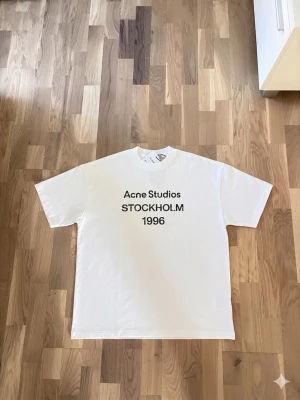 Vit Acne Studios t-shirt Stockholm - Vit t-shirt från Acne Studios med svart text 'Acne Studios STOCKHOLM 1996' på bröstet. Klassisk rak passform och rund halsringning. Tillverkad i mjuk bomull, perfekt för en clean och stilren look.