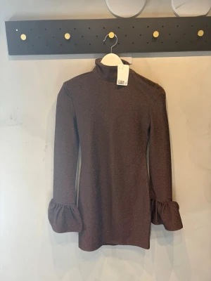 Brun glittrig polotopp med volangärm - Snygg brun polotopp med glittrigt tyg och långa ärmar som avslutas med bred volang. Perfekt för dig som vill sticka ut lite extra med en trendig och elegant look. Polokrage och figurnära passform med halvrygg dragkedja.