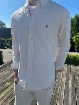 Vit skjorta från Ralph Lauren - Klassisk vit skjorta från Ralph Lauren med button-down krage och broderad logga på bröstet. Skjortan har långa ärmar, knappar framtill och är tillverkad i mjuk bomull. Perfekt för en clean och stilren look.
