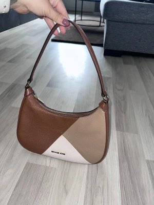 Brun och beige handväska Michael Kors - Snygg axelväska från Michael Kors i brunt, beige och vitt läder med patchwork-design. Väskan har en mjukt rundad form och guldfärgad dragkedja. Perfekt accessoar för dig som gillar stilrena detaljer och klassisk look.