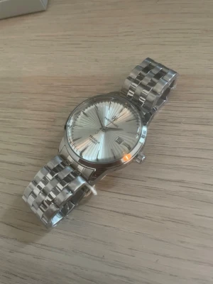 Silverfärgad klocka från Pablo Raez - Stilren automatisk klocka från San Martin med silverfärgad urtavla och matchande metallarmband. Klockan har datumvisning och klassisk rund form. Perfekt accessoar för dig som gillar tidlös design och metalliska detaljer.