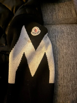 Svart Moncler cardiganjacka med huva - Snygg svart cardiganjacka från Moncler med vadderad kropp och stickade ärmar. Ena ärmen har ett vitt zigzag-mönster och Moncler-logga. Jackan har dragkedja framtill, fickor och en skön huva. Perfekt för dig som vill ha en trendig och bekväm look.