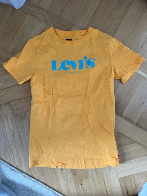 Gul Levi's t-shirt med blå logga - Snygg gul t-shirt från Levi's med klassisk blå logga tryckt på bröstet. Tillverkad i mjuk bomull och har en avslappnad passform med korta ärmar och rund hals. Perfekt för dig som gillar färg och vill sticka ut lite extra.