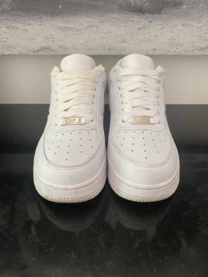 Nike Air Force 1 vita sneakers - Klassiska Nike Air Force 1 sneakers i helvitt läder med perforerad tå, vit sula och ikonisk Swoosh på sidan. Skorna har lågt skaft och vita skosnören. Perfekta för en clean och stilren look.