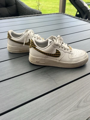 Nike Air Force 1 med rutiga detaljer - Säljer ett par vita Nike Air Force 1 sneakers med unika gul- och svart-rutiga detaljer på swoosh och häl. Klassisk låg modell med perforerad tåbox och vita skosnören. Tillverkade i läder med gummisula. Perfekta för dig som vill sticka ut med en personlig touch. Köpta på Nike store