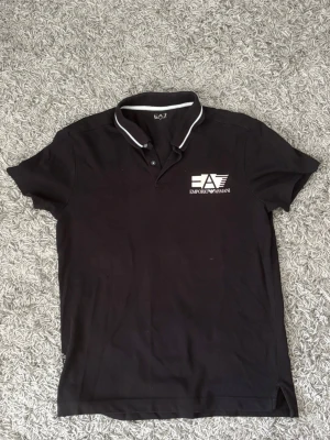 Svart pikétröja från Emporio Armani - Snygg svart pikétröja från Emporio Armani EA7 med vit logga på bröstet och vit detalj på kragen. Klassisk pikékrage med knappar och korta ärmar. Perfekt för en stilren och sportig look.