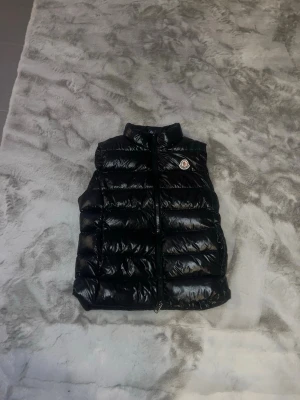Svart dunväst från Moncler - Snygg svart dunväst från Moncler med glansig finish och hög krage. Västen har dragkedja framtill och det klassiska Moncler-logotypen på bröstet. Perfekt för lager-på-lager och ger en clean streetwear-look. Pris kan diskuteras vid snabb affär.