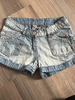 Blå jeansshorts från Girl Star - Säljer ett par ljusblå jeansshorts från Girl Star med snygga detaljer som stora fickor fram och bak, knappstängning och uppvikta benslut. Perfekta till sommaren och har en klassisk denimlook som alltid funkar.