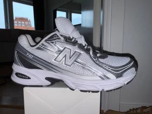 New Balance 740 vita sneakers - Snygga New Balance 740 sneakers i vitt och silver med mesh- och syntetmaterial. Skorna har den klassiska N-loggan på sidan, vita skosnören och en chunky sula med ABZORB-dämpning. Perfekta för dig som gillar sportig och trendig stil.