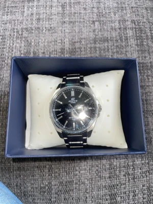 Casio Edifice svart/silver klocka - Stilren Casio Edifice klocka med svart urtavla och datumvisning. Armbandet är i blankt silverfärgat metall och klockan har en klassisk rund form. Perfekt för dig som gillar en tidlös och elegant accessoar.