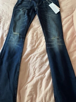 Mörkblå bootcut jeans med slitningar - Snygga mörkblå bootcut jeans med slitningar på låren och knäna. Jeansen har en klassisk femficksmodell och coola slitna detaljer framtill. Materialet är denim med en lätt stretch för skön passform. Perfekta för en trendig och avslappnad look.