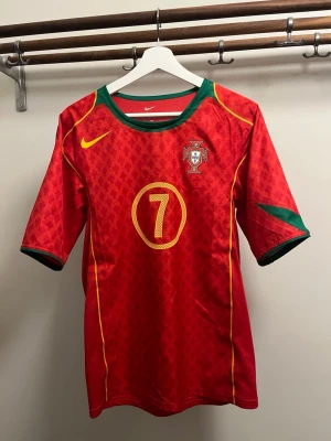 Portugal Nike fotbollströja Figo #7 - Röd Portugal fotbollströja från Nike med gula detaljer, gröna inslag vid hals och ärmar samt nummer 7 och namnet Figo på ryggen. Tröjan har korta ärmar, broderad landslagssköld och är tillverkad i lätt, ventilerande material.