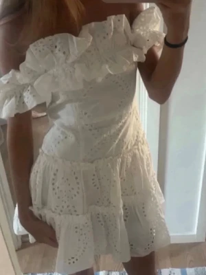 Vit offshoulder klänning med volang - Söt vit kortklänning med offshoulder och volangdetaljer. Klänningen har broderade hålmönster och en figurnära passform upptill med utsvängd kjol. Perfekt för sommarens alla tillfällen.
