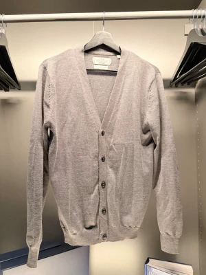 These Glory Days  - Stilren ljusgrå cardigan med v-ringning och knäppning framtill. Tillverkad i ett tunt, mjukt material som passar perfekt för lager-på-lager. Långa ärmar och ribbade muddar ger en klassisk look.
