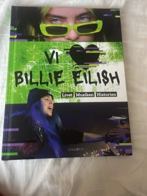 Vi ♥ Billie Eilish - Livet Musiken Historien - En färgstark bok om Billie Eilish med coolt omslag i grönt, svart och vitt. Innehåller fakta, foton, quiz och citat om Billies liv, musik och karriär. Perfekt för fans som vill veta allt om stjärnan och följa hennes resa till toppen.