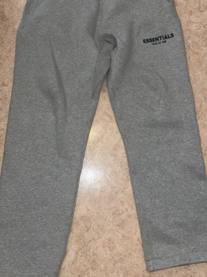 Grå Essentials mjukisbyxor - Säljer ett par grå mjukisbyxor från Essentials Fear of God. Byxorna har resår och dragsko i midjan samt sidofickor. Tillverkade i mjukt material som känns skönt mot huden. Perfekta för chill dagar eller när du vill ha en avslappnad stil.